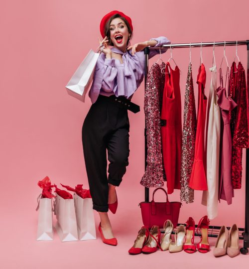 Woman in black trousers and purple blouse laughs, leaning on stand with elegant clothes on pink background